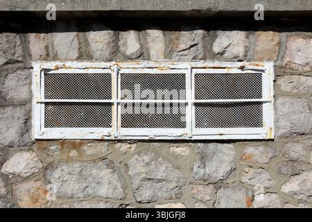 A rectangular window with a rusted metal grille and mesh is set in a rough stone wall, showing signs of age and weathering, with sunlight casting Stock Photo