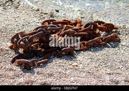 A thick, rusty metal chain lies coiled on a pebbled shore at the water’s edge, its corroded links contrasting with the light stones, evoking strength Stock Photo