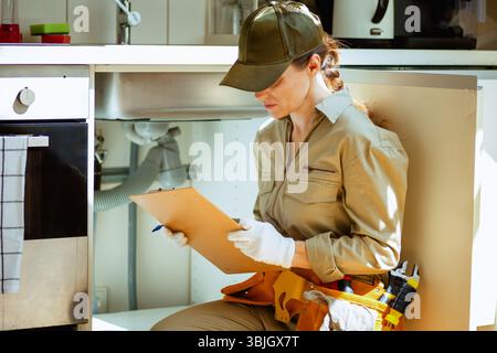 A focused plumber inspects plumbing under a kitchen sink, ensuring everything is working correctly. Stock Photo
