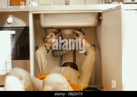 A plumber lies on his back, working on pipes under a kitchen sink, using a light. The scene emphasizes skilled home maintenance and problem-solving. Stock Photo