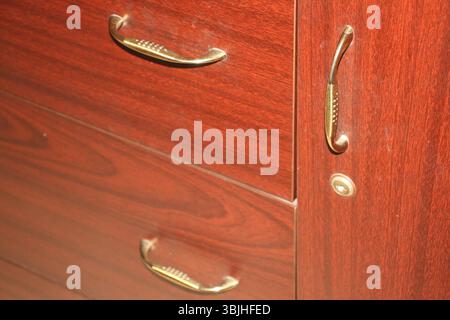 A close-up view of a wooden cabinet made of polished dark reddish-brown wood. The cabinet has three metallic brass handles with curved design. Stock Photo