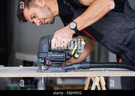 Worker in workwear using handheld cordless electric jigsaw to cut wooden board in garage. Skilled carpenter working in workshop. Man doing DIY home re Stock Photo