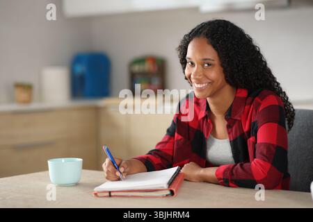 Happy black student taking notes looks at you in the kitchen at home Stock Photo