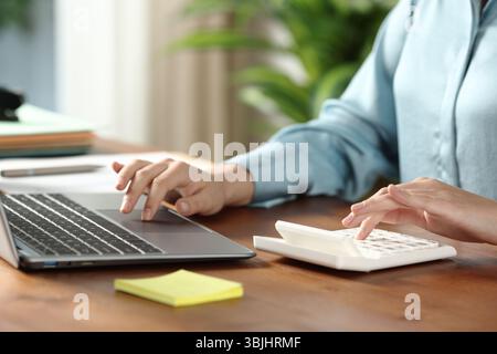 Close up portrait of an office worker calculating with calculator and browsing on laptop on wooden table Stock Photo