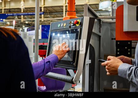 Industrial worker operating CNC machine control panel with touchscreen interface at manufacturing exhibition. Modern automated equipment with digital Stock Photo