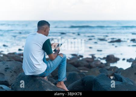 Barefoot man sits on a coastal rock, focused on his smartphone with a calm sea in the background. Stock Photo