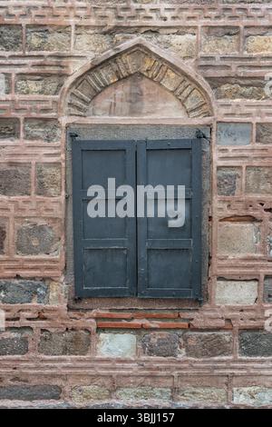 Old stone and brick wall with arched window and closed dark blue metal shutters. Weathered architectural detail. Stock Photo