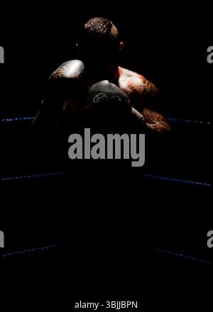 A male boxer with tattoos, his face obscured by shadow, stands in a defensive stance in a dimly lit boxing ring. The focus is on his upper body Stock Photo
