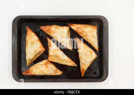 Golden brown, triangular pastries on a baking tray arranged in a pattern against a plain white background Stock Photo