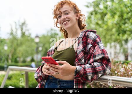 Smiling redhead woman with tattoos and piercings using mobile phone and holding coffee cup while leaning on railing in a city park Stock Photo