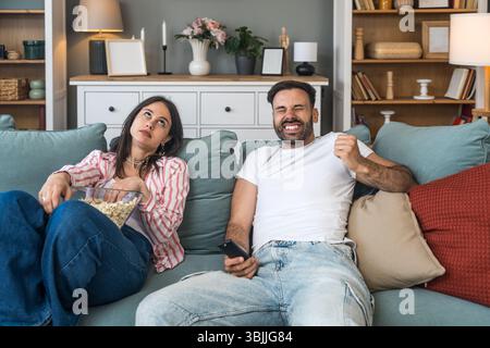 Young couple sitting together on sofa at home during first date night, watching sports or movie on TV, man fully engaged and excited, while woman look Stock Photo