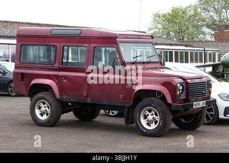 A red Land Rover Defender parked on a tarmac, surrounded by other vehicles with a brick building and trees in the background Stock Photo