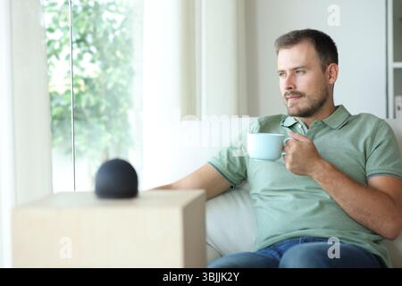 Relaxed man with smart speaker drinking coffee sitting on a couch at home Stock Photo
