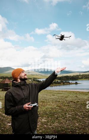 Smiling man reaching for a drone in mid-air above a lake in Zlatibor, Serbia. Beautiful spring mountains and blue sky behind. Travel blogger making Stock Photo
