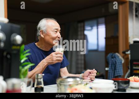 In a small Japanese apartment, a Japanese man in his late 70s relaxes in T-shirt pajamas. He sits alone in the evening, sipping whiskey and enjoying a Stock Photo