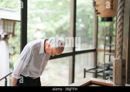 In rural Japan, a Japanese man in his late 70s visits a countryside shrine surrounded by lush green trees. Wearing a white shirt and hat, he prays qui Stock Photo