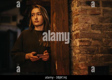 Young woman holding smart phone standing by a rustic brick wall indoors Stock Photo