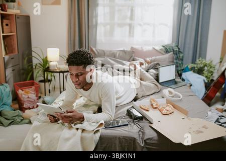 Young Man Lying on Bed Using Tablet in Cluttered Room Stock Photo