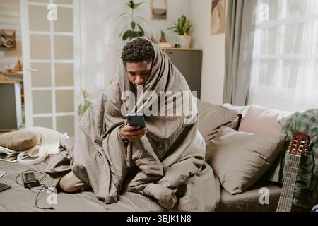 Man Wrapped in Blanket Using Smartphone Indoors Stock Photo