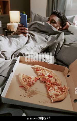 Relaxing in Bed Enjoying Pizza While Using Mobile Device Stock Photo