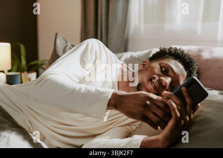 Smiling Young Man Relaxing in Bed With Smartphone Stock Photo