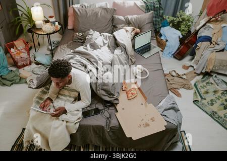 Working on Tablet While Lying on Bed Surrounded by Clutter Stock Photo