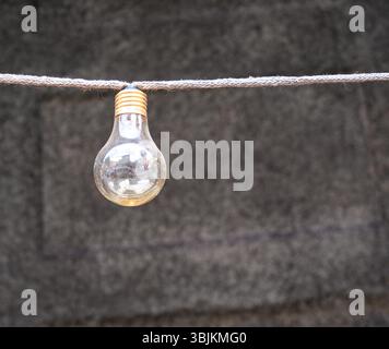 A single light bulb hanging from a string against a textured gray background. The bulb is clear with a metallic base, showcasing its vintage design. Stock Photo