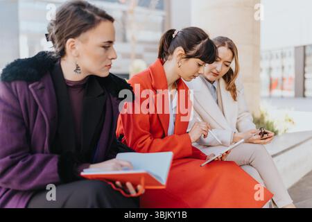 Three businesswomen collaborating outdoors and discussing ideas using a notebook and tablet Stock Photo
