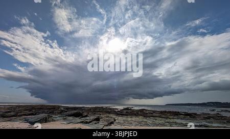 A massive cloud formation builds over a rocky coastline with visible sunlight breaking through, capturing a moment before a storm. Stock Photo