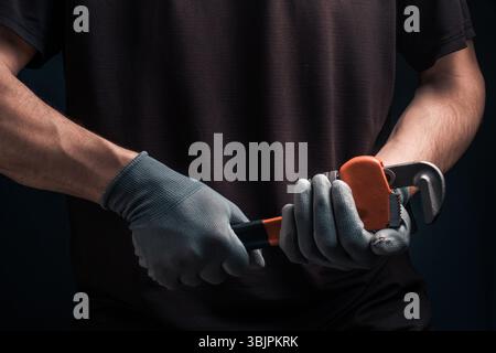 Plumbers hands in protective gloves holding pipe wrench on dark background Stock Photo