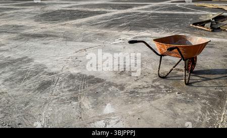 Old orange construction wheelbarrow on textured concrete ground at urban building site, photographed in natural sunlight. No people. Stock Photo