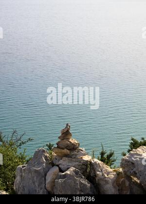 A stack of brown pebbles framed by greenery and rocks on the coast, with the shimmering turquoise sea and soft waves behind. Stock Photo
