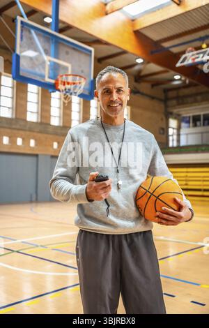 Mature basketball coach holding a ball and stopwatch, smiling confidently in an empty gymnasium, embodying passion for the sport Stock Photo