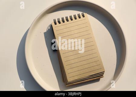 Minimalist top-down still life. Brown empty spiral notebook mockup with lined pages placed on ceramic plate, soft natural light. Creative, conceptual Stock Photo
