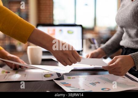 Closeup of hands of female accountant exchanging documents, collaborating at office desk in analyzing company expenses report. Selective focus of individuals holding papers over the table. Stock Photo