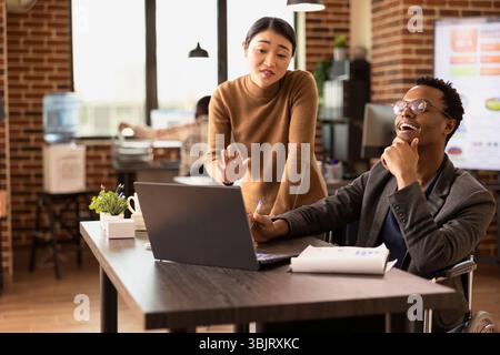 Smiling businessman listening as his asian coworker explains market trends using digital chart on laptop. Diverse team brainstorming strategy ideas, reviewing financial analytics on an office desk. Stock Photo