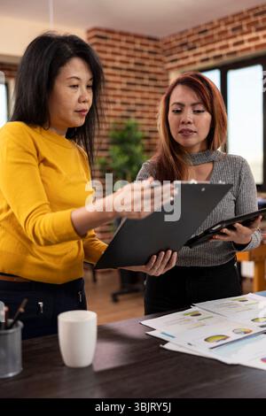 Asian businesswomen collaborate on strategy planning, comparing ideas with a clipboard and tablet in a modern workspace. Two female colleagues stand in office, reviewing each others project notes. Stock Photo