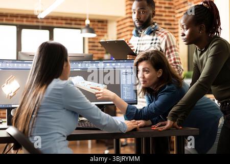 Young multiethnic technicians designing a prototype for the manufacturing industry. Diverse engineers collaborating around the table, developing a mechanical clamp using CAD software. Stock Photo