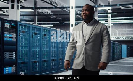Smiling engineer in server hub monitoring data center energy consumption across components. Happy IT expert walking in data center, making sure rigs sensors are functioning optimally Stock Photo
