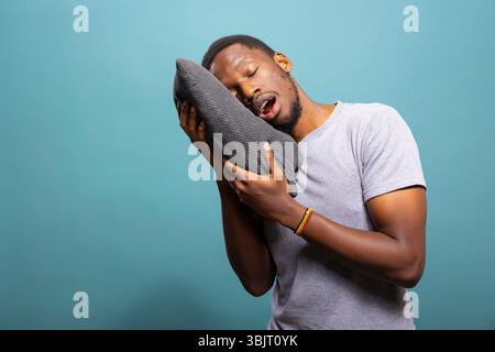 Tired young man with mouth open and eyes closed, resting his head on a pillow, showing exhaustion. Black male model pretending to sleep on a gray cushion while standing against a blue background. Stock Photo
