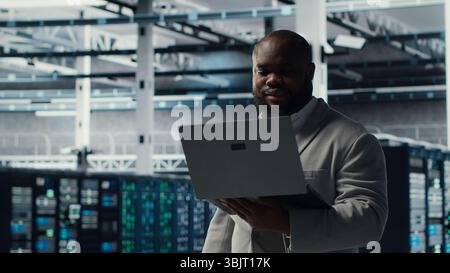 Server room worker auditing systems for vulnerabilities using software on laptop. Engineer walking in data center doing backups and recovery protocols using notebook, camera B Stock Photo