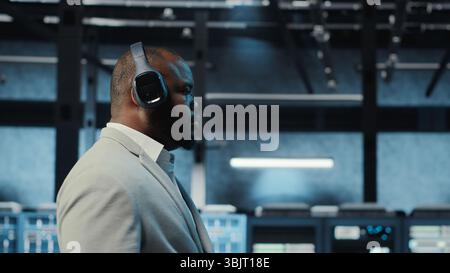 Happy manager listening music, walking in data center with blinking lights on rigs showing system activity. Cheerful man wearing headphones overseeing dimly lit server farm, camera B Stock Photo