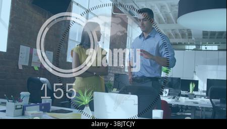 Discussing blue-shirted man and yellow-dressed woman using laptop in open office with graphics Stock Photo