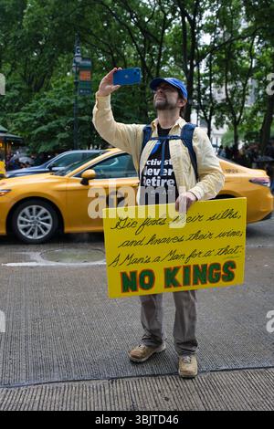 A man in a yellow shirt and blue cap holds a yellow sign with a quote and the words "NO KINGS" in green. He's taking a selfie with a smartphone in a c Stock Photo