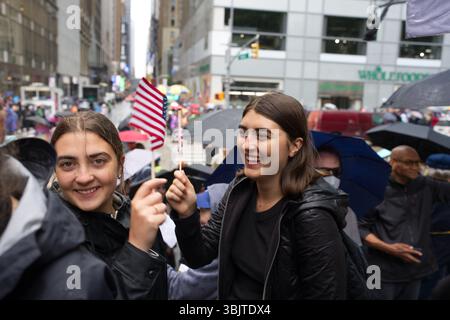 Two young women hold small American flags on a rainy city street, amid a crowd of people with umbrellas.  The women are smiling, and the background sh Stock Photo