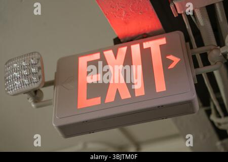Close-up of a red EXIT sign with built-in emergency light, mounted on a ceiling near electrical conduit. Symbol of safety and emergency evacuation. Stock Photo
