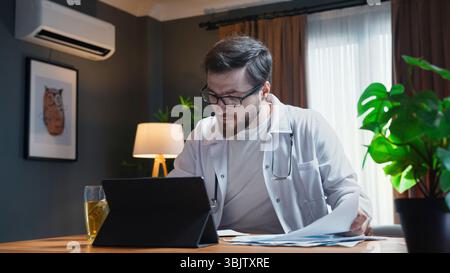 Male doctor in white coat and glasses concentrates on tablet while reviewing medical documents in a cozy home office setting. Stock Photo