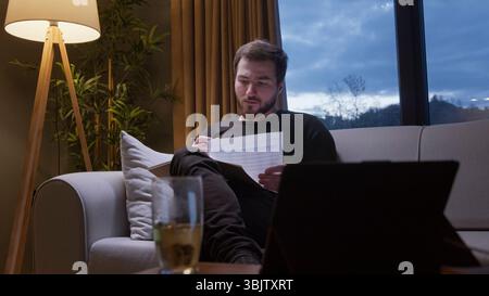 Focused man sits on sofa at dusk reading printed charts, with tea and laptop nearby, suggesting remote work or study. Stock Photo