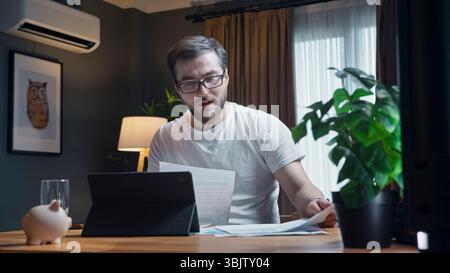 Young man in white t-shirt studies paperwork at desk while working remotely on laptop in a cozy home office environment. Stock Photo