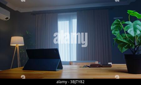 Minimalist workspace featuring a wooden desk, closed tablet, and soft daylight through curtains in a quiet modern room. Stock Photo